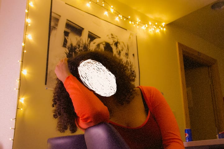 A photo of a woman sitting in a dimly lit room. Her face is scribbled out with white ink
